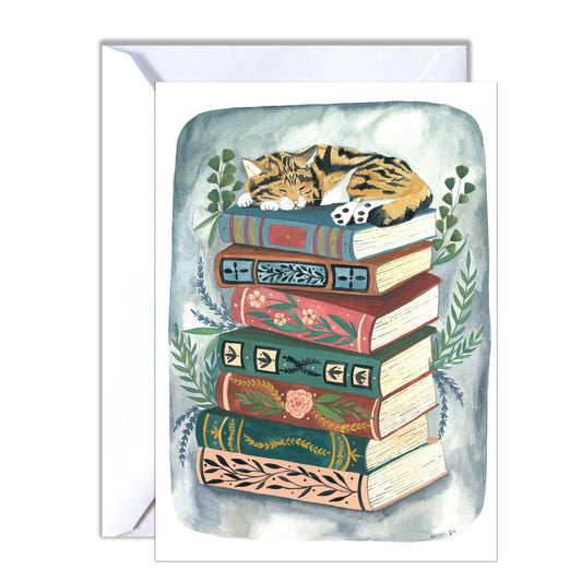 Greeting card featuring watercolor illustration of a tabby cat sleeping atop a stack of vintage books. The books have ornate designs and floral accents in muted tones of green, pink, and red.  The cat is light brown and dark brown tabby. Greenery surrounds the base of the book stack. The card is suitable for cat lovers and bookworms.